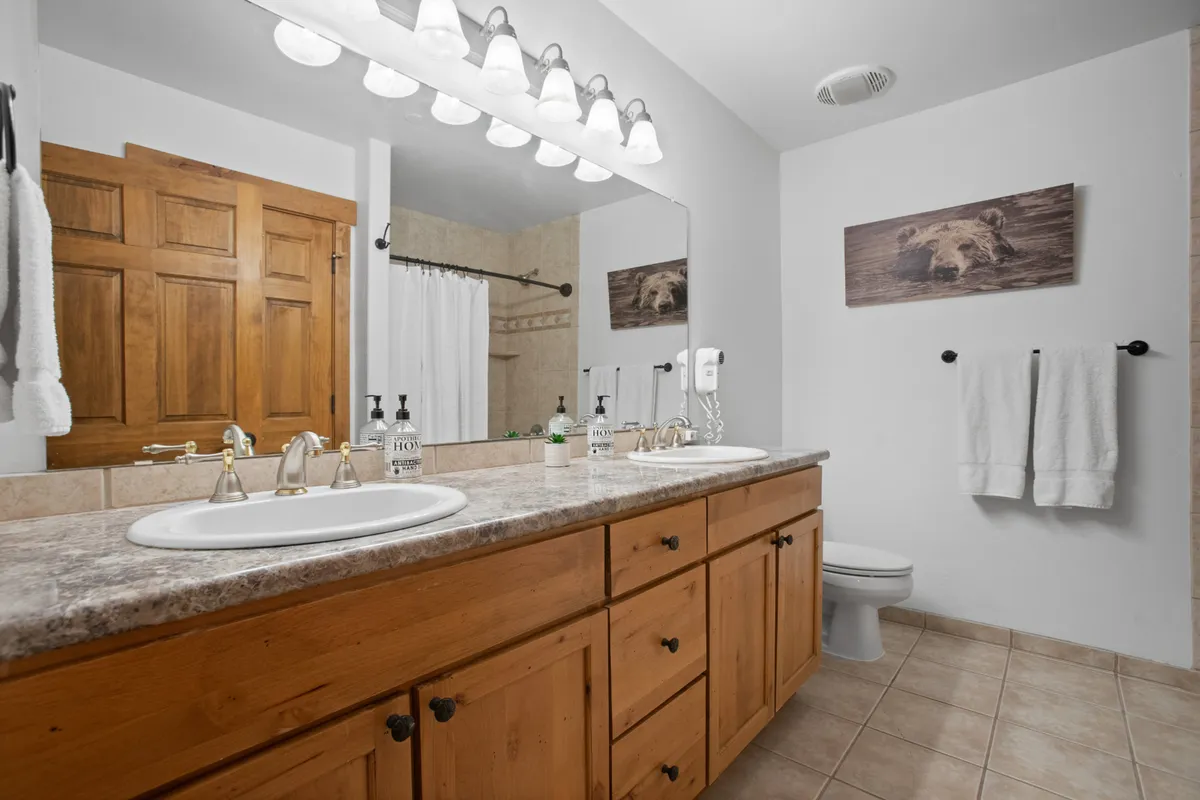 A spacious bathroom featuring a double sink with elegant golden fixtures, a wide mirror, wooden cabinetry, and distinctive bear-themed artwork. The design is fresh and tranquil, accommodating multiple guests comfortably.