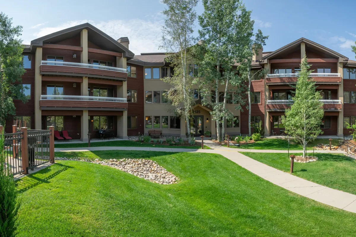 A welcoming exterior shot featuring balconies overlooking a green lawn, a meandering pathway, and mature trees. The building's design blends with the mountain surroundings, and the spaces invite guests to relax outdoors or enjoy a stroll.