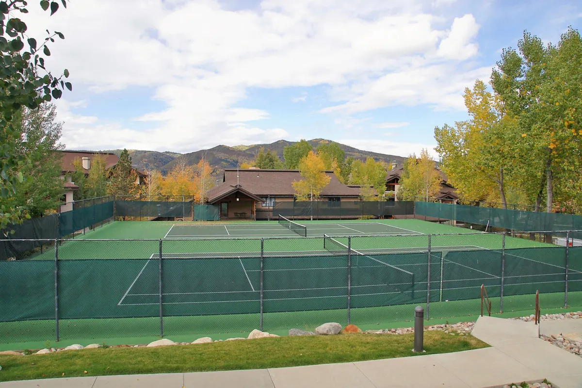 A vibrant exterior view of tennis courts set amidst lush green trees with a stunning mountain backdrop. The open sky and surrounding natural scenery provide an inviting environment for outdoor recreation.