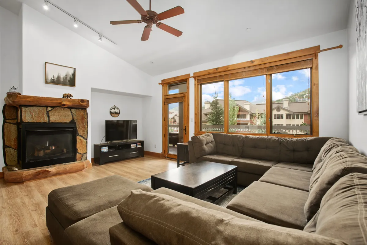 The image showcases a stylish, spacious living area anchored by a stone fireplace, with a plush sectional sofa arranged for relaxation. Large windows flood the space with natural light and reveal picturesque views of the ski mountain. The inviting atmosphere is perfect for unwinding after a day on the slopes or gathering with family and friends.
