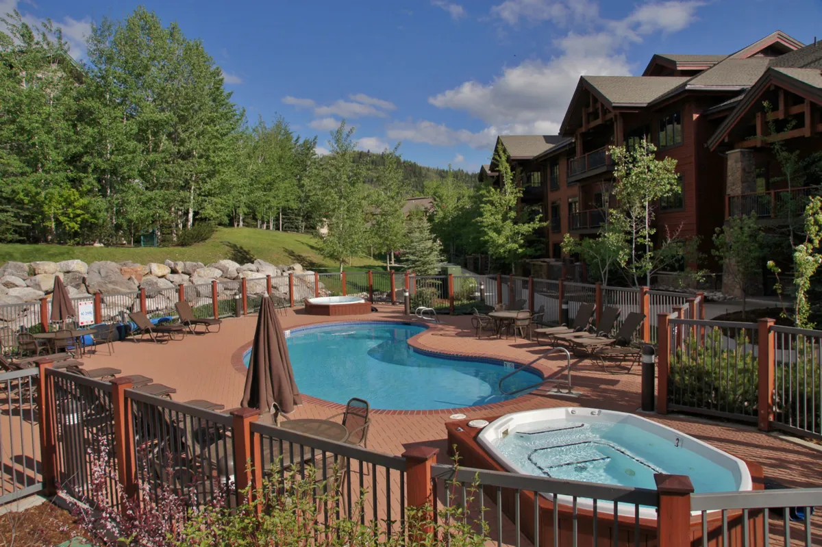 A vibrant outdoor scene featuring a sparkling swimming pool and adjacent hot tub surrounded by lush greenery and inviting lounge chairs. The setting is perfect for group gatherings and relaxation, showcasing an atmosphere that caters to both summer and winter enjoyment.