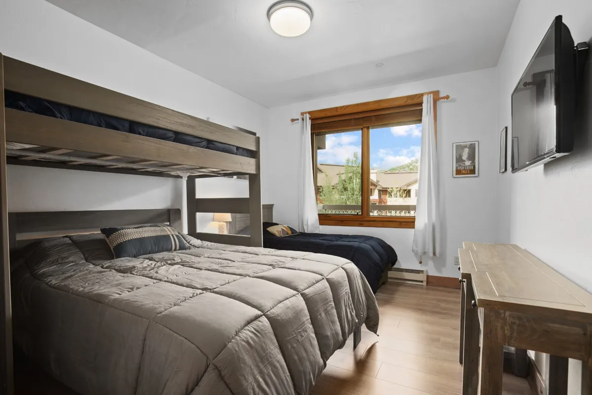 A flexible family bedroom with a twin-over-queen bunk bed and an additional bed, bright window, and a wall-mounted TV. This inviting space is functional and perfect for groups or families, offering flexible sleeping arrangements and entertainment.