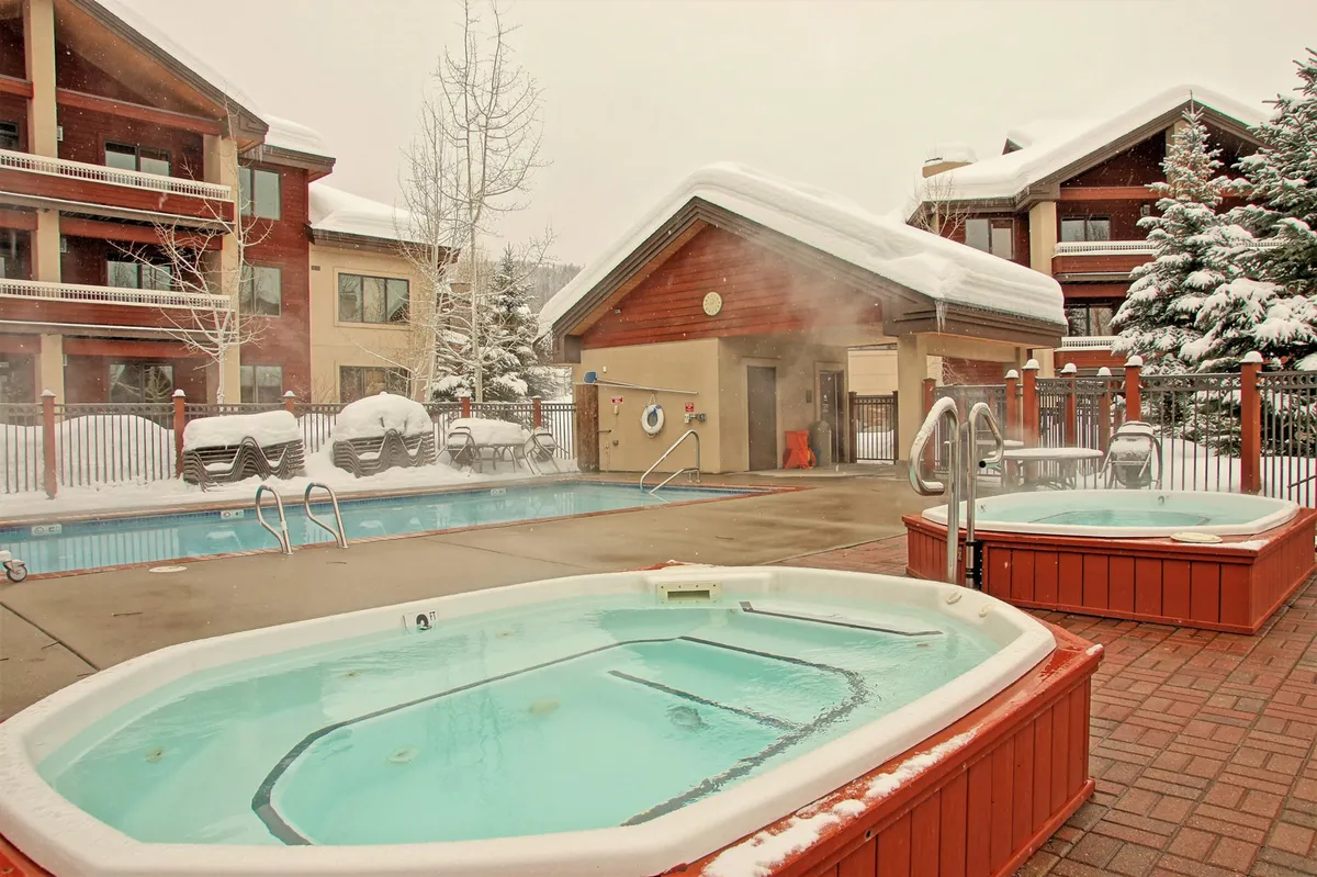 This image captures a scenic outdoor pool area with multiple hot tubs, snow-covered surroundings, and inviting wooden architecture. People are seen enjoying hot tub soaks while snow gently falls, conveying relaxation and luxury in a mountain resort setting.