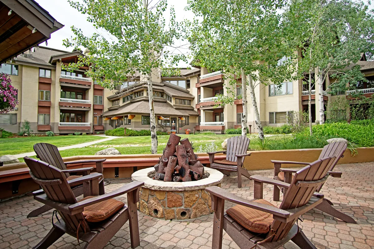 An exterior view of a cozy fire pit area with wooden chairs arranged around the fire, set within a landscaped garden and backed by the building's facade. The scene evokes a sense of warmth, social connection, and relaxation during the evening or after outdoor adventures.