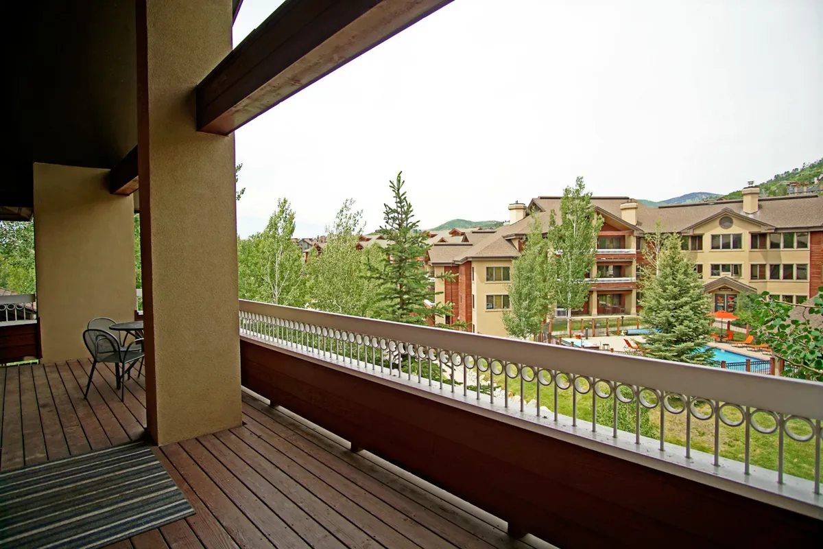 A peaceful balcony scene featuring a wooden deck and railing surrounded by lush trees, with distant buildings in view. The space is set for relaxation, inviting guests to enjoy morning coffee or a sunset drink in the fresh mountain air.