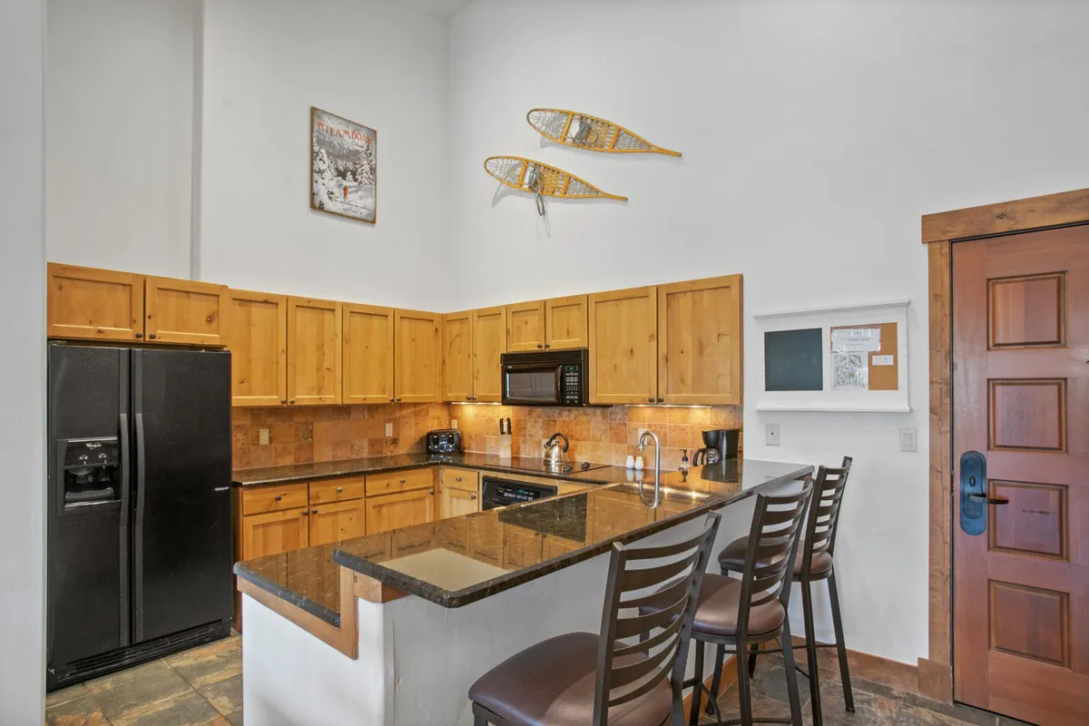 A kitchen scene featuring gleaming granite countertops, wooden cabinetry, black appliances, and the entry door in view. The space is both practical and social, inviting guests to cook, snack, and mingle.